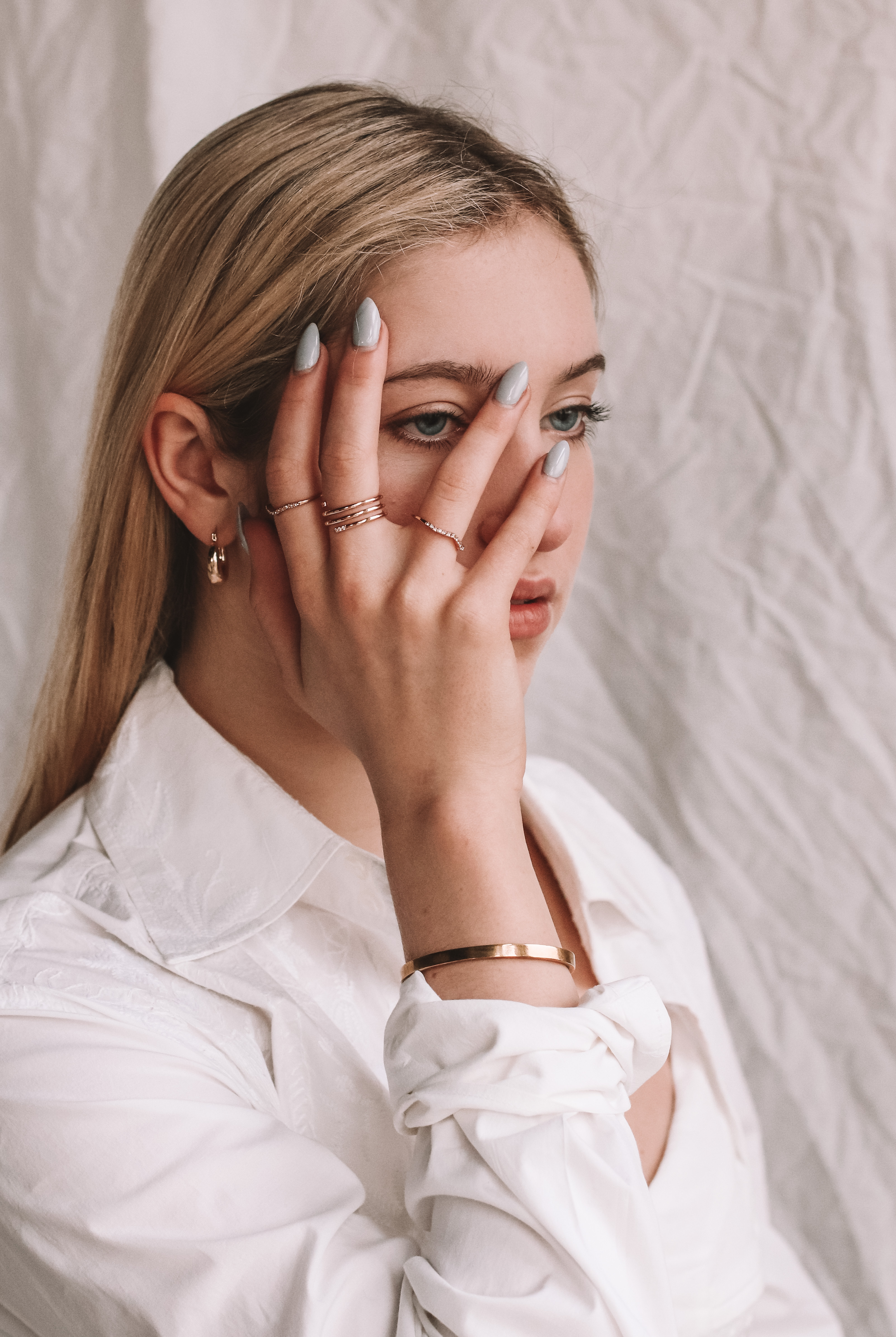 editorial photo of a girl with blonde hair wearing a white button-down and gold jewelry framing her face with her hands